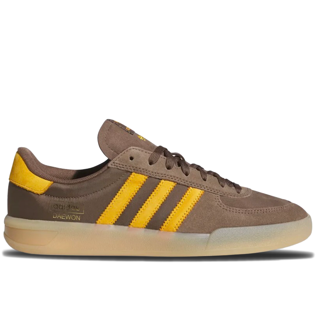 A brown suede and leather low-top sneaker with three yellow stripes, a yellow heel tab, and a translucent beige gum sole, shown in a side profile on a white background.