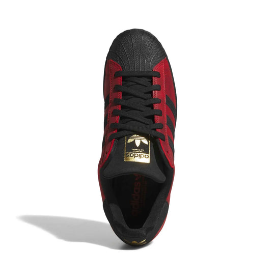 An Adidas Superstar sneaker in black and red, featuring a metallic gold logo plate on the tongue, photographed from a top-down angle on a white background.