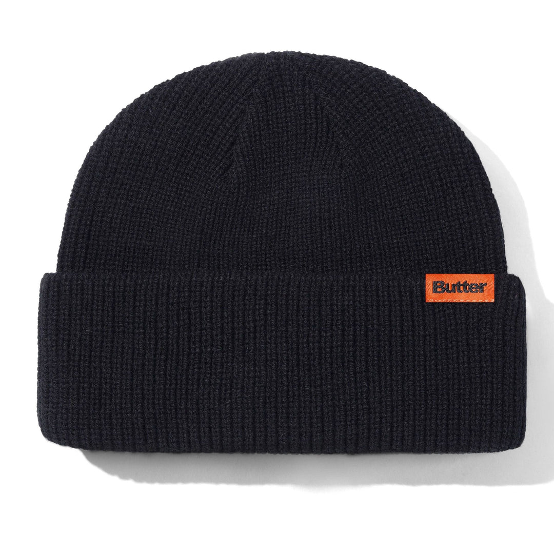 A black ribbed knit beanie hat with a small orange rectangular Butter logo patch on the front cuff, shown on a white background with a soft shadow.