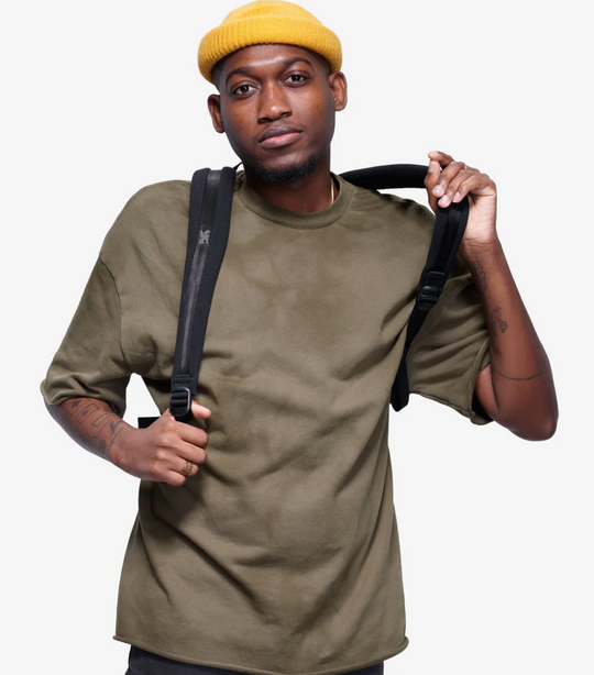 Man wearing a green t-shirt and yellow beanie holding a black bag against a white background