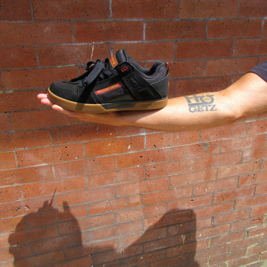 A black skate shoe with a tan gum sole and orange accent stripes, held in a person&
