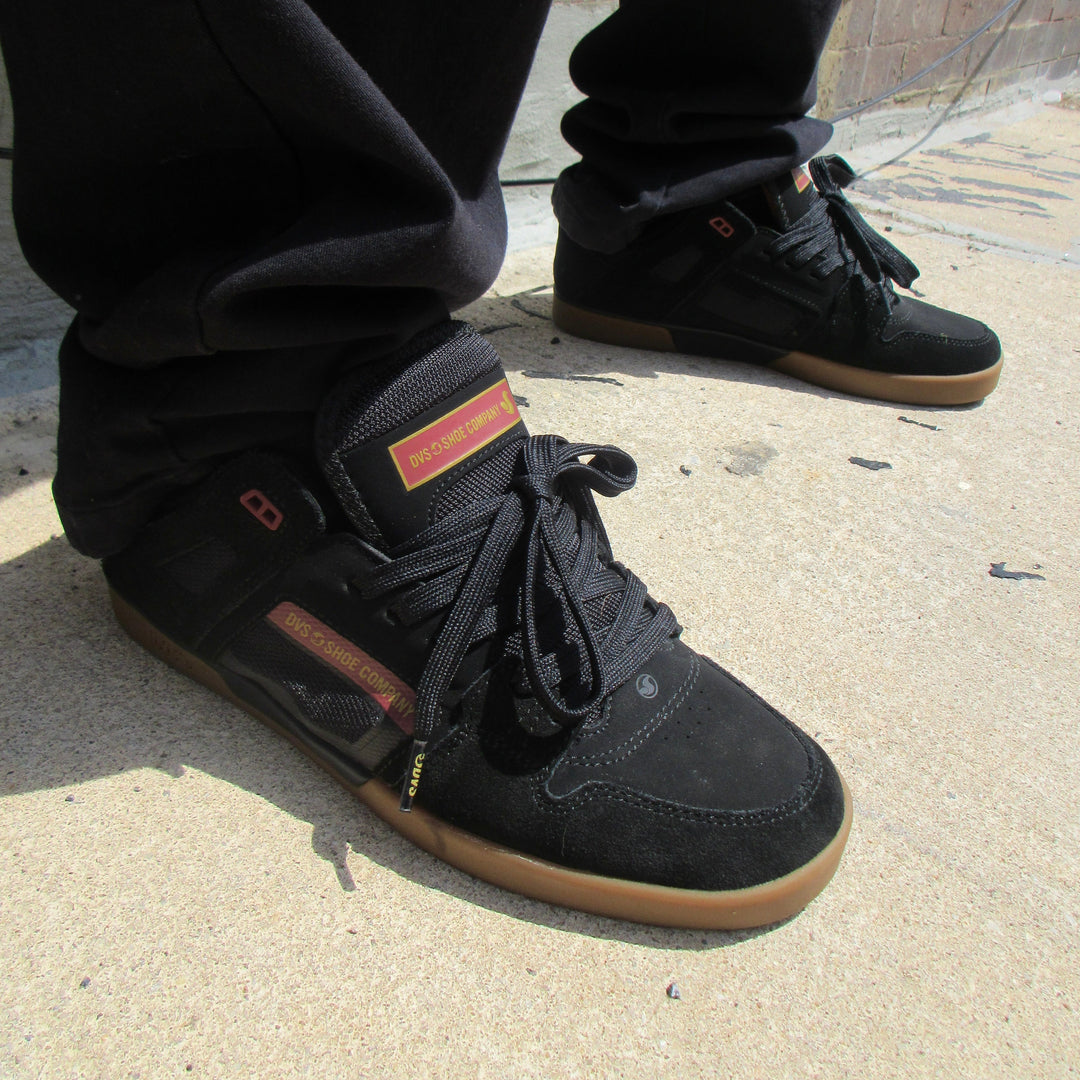 A person wearing black high-top skate shoes with gum soles, paired with black pants, standing on a concrete ground.