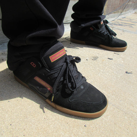 A person wearing black high-top skate shoes with gum soles, paired with black pants, standing on a concrete ground.