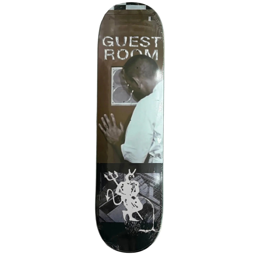 A skateboard deck with a graphic print featuring the text 'GUEST ROOM' and a photo of a man, displayed on a textured gray sheet of paper between wooden boards.