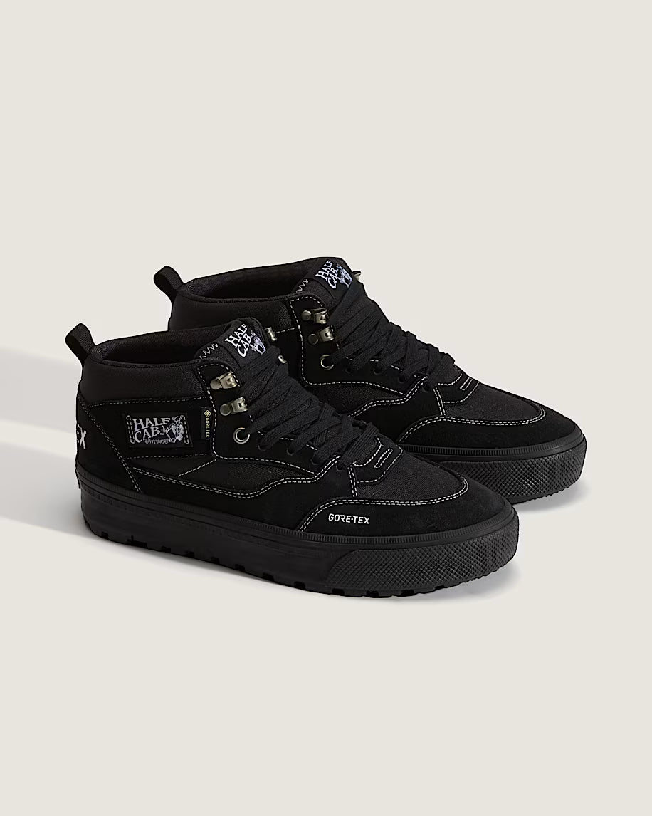 A pair of black mid-top sneakers with white contrast stitching, white GORE-TEX branding on the side, and white embroidered details on the tongue and heel, set against a light beige background.