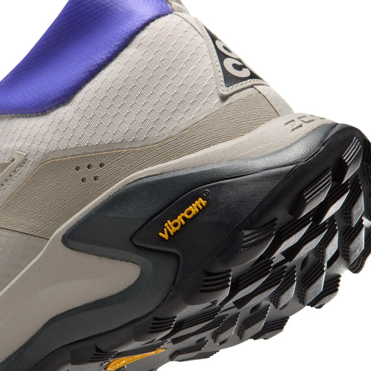 Close-up of a trail running shoe showing the midfoot and outsole, featuring a beige and gray upper, a purple collar, and a black Vibram sole.
