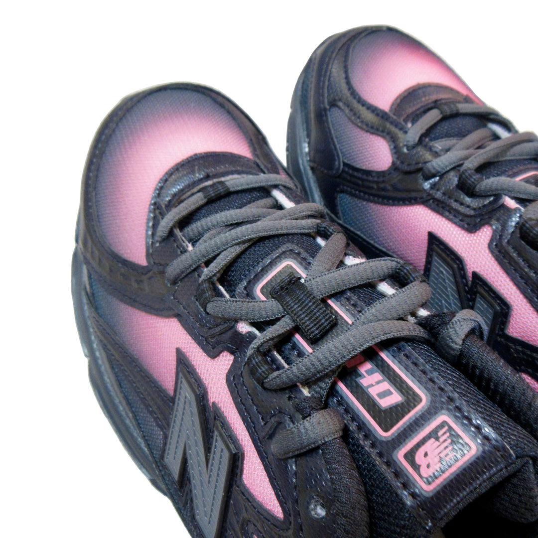 A close-up of a pair of New Balance 990v3 sneakers in black and pink, showing the side profile, laces, and the New Balance logo on the tongue and side.