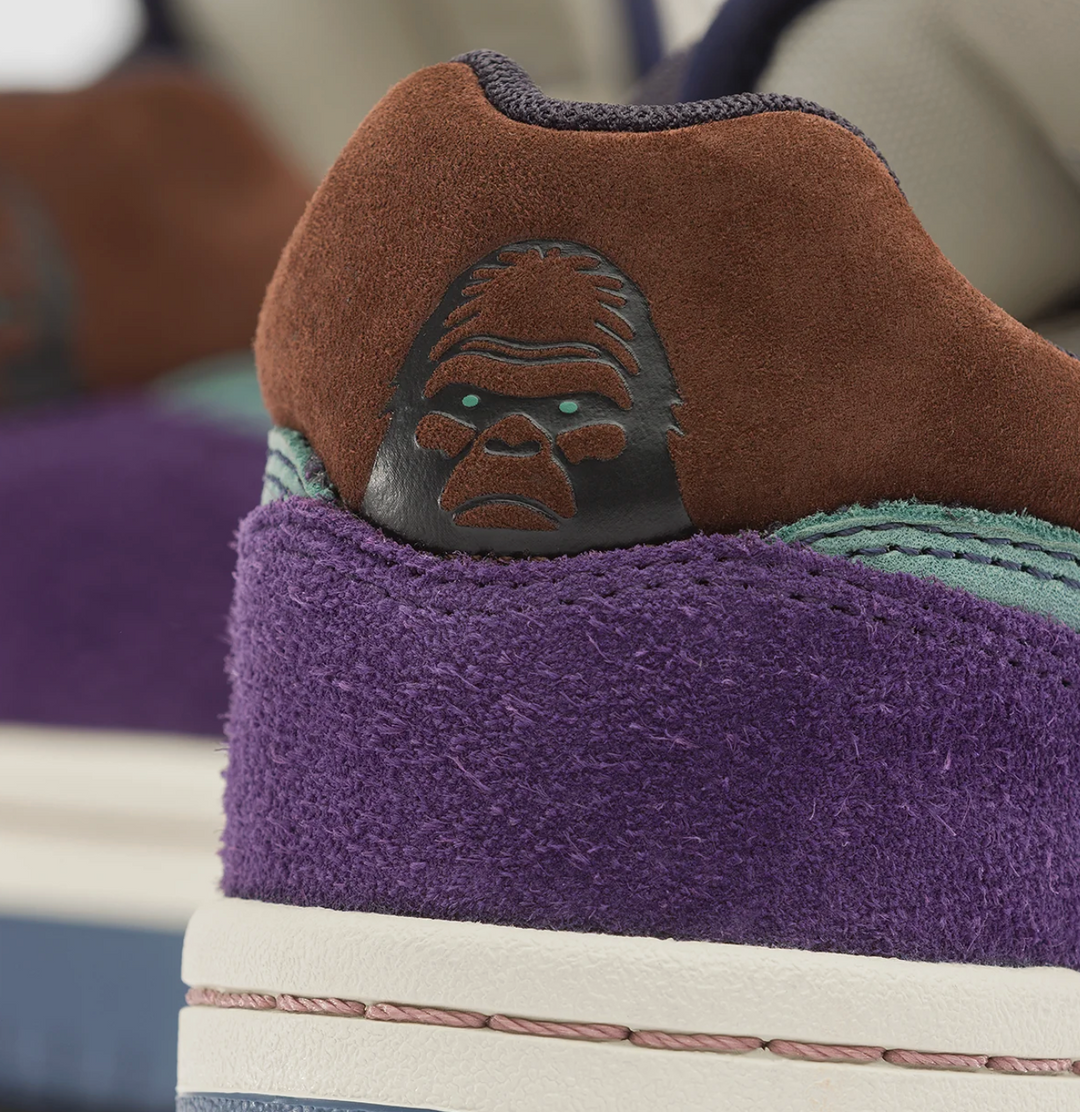 Close-up of a sneaker showing a purple suede heel counter with an embossed gorilla face logo on a brown suede collar, a white midsole, and a teal eyelet area.