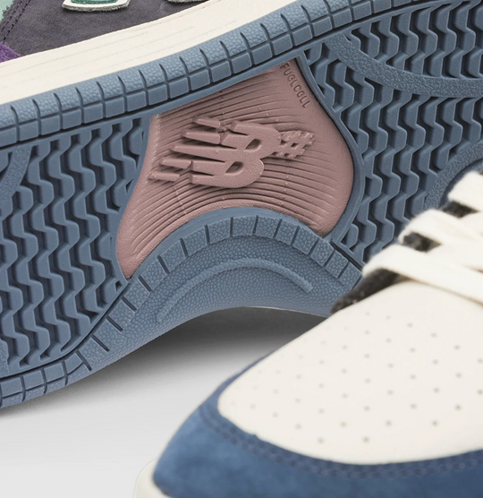 Close-up of a New Balance sneaker showing the textured blue-gray outsole with a dusty pink heel insert and the embossed New Balance &