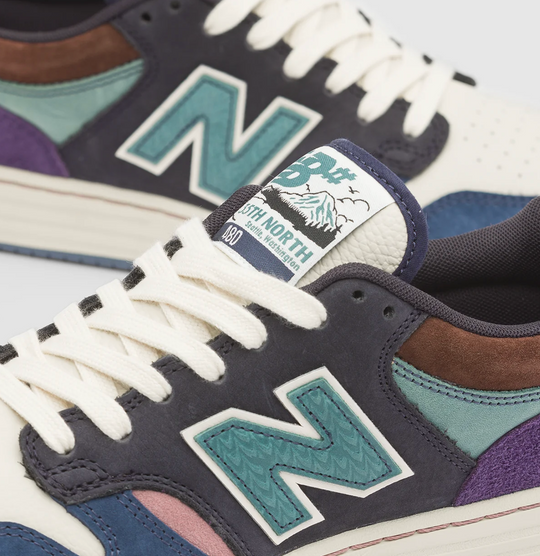 Close-up of a New Balance sneaker in dark navy suede with teal, purple, brown, and pink accents, cream laces, and a visible tongue label reading &