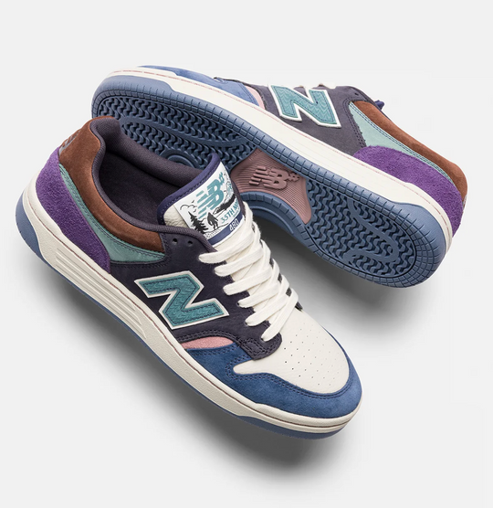 A pair of New Balance 990v5 sneakers in a multi-color colorway featuring purple suede, navy suede, brown leather, teal overlays, and white leather. One shoe is positioned to show the side profile with the &