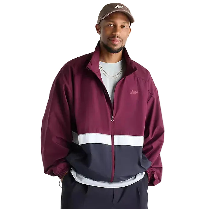 A man wearing a New Balance colorblock windbreaker jacket in burgundy, white, and navy, paired with a brown cap, white t-shirt, and navy pants, against a plain white studio background.