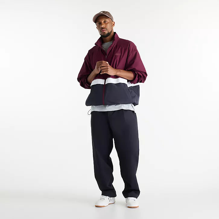 A man modeling a colorblock windbreaker jacket in burgundy, navy, and white, paired with dark navy pants, white sneakers, and a brown cap, shot against a seamless white studio background.