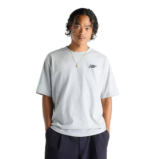 A man wearing a light gray heather crewneck t-shirt with a small black New Balance logo on the left chest, paired with dark navy pants, against a plain white studio background.