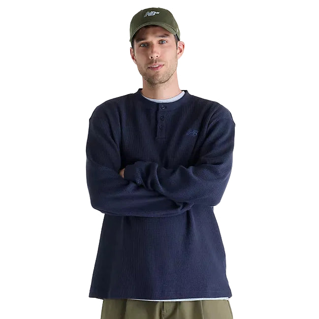 A man wearing a navy blue ribbed henley shirt, olive green New Balance cap, and olive green pants, standing with arms crossed against a plain white background.