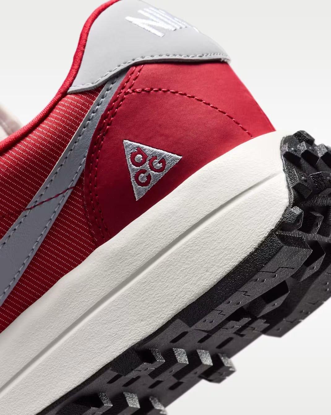 Close-up of a red and gray sneaker showing the heel, side stitching, triangular logo patch, white midsole, and black treaded outsole.