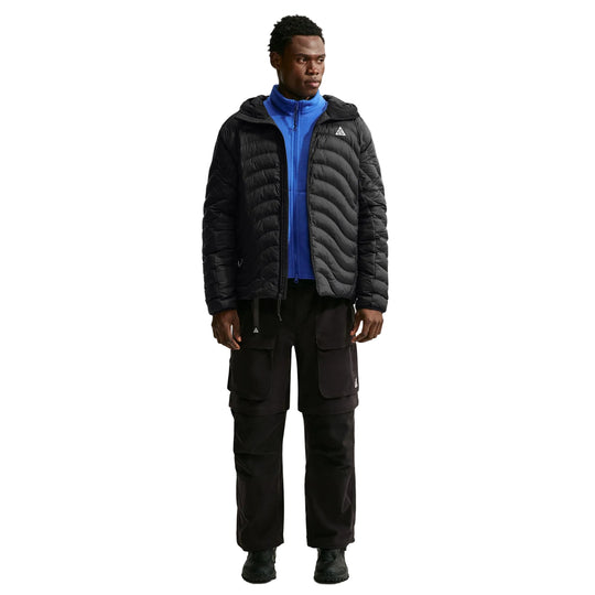 A man modeling a black ACG puffer jacket with a wavy quilted texture, worn over a bright blue zip-up fleece and black cargo pants, against a plain light gray studio background.