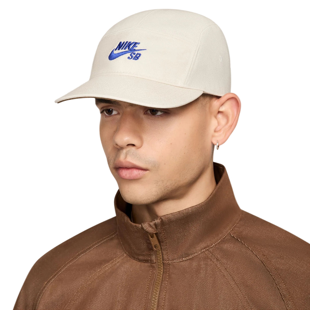 A man wearing a beige Nike SB baseball cap with a blue embroidered logo, paired with a brown zip-up jacket, against a light gray studio background.