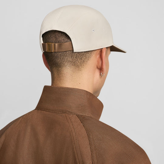 Rear view of a man wearing a light beige baseball cap with a brown adjustable strap and a brown collared jacket against a light gray background.