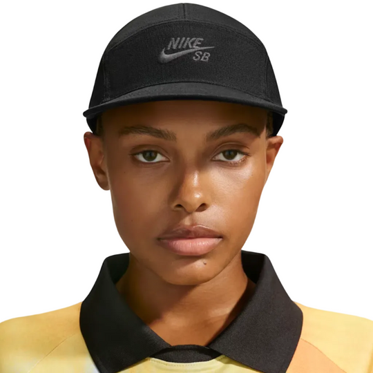 A close-up of a model wearing a black Nike SB baseball cap and a yellow polo shirt with a black collar, set against a clean white studio background.