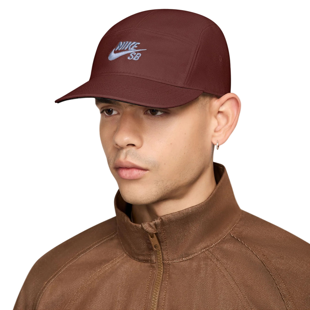 A man wearing a dark maroon Nike SB cap and a brown zip-up jacket, shown in a close-up profile against a light gray background.