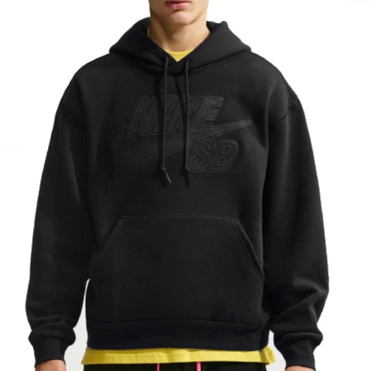 A man wearing a black Nike SB hoodie with a tonal black logo on the chest, layered over a yellow t-shirt and black sweatpants with pink drawstrings, against a plain white background.
