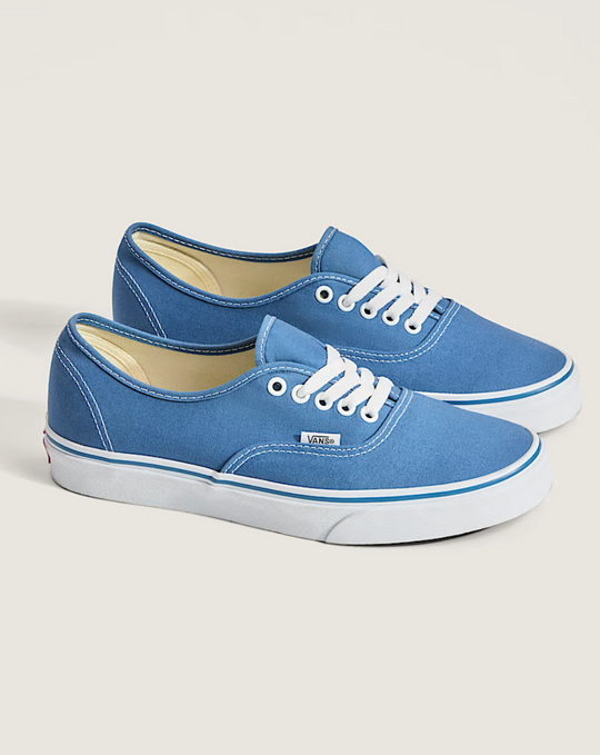 A pair of Vans Authentic low-top canvas sneakers in a medium blue colorway, featuring white laces, white rubber midsoles with blue stripes, and a light beige interior lining, set against a solid light beige background.